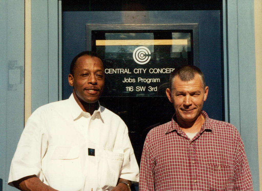 Two men standing in front of CCC jobs program sign in the 90s