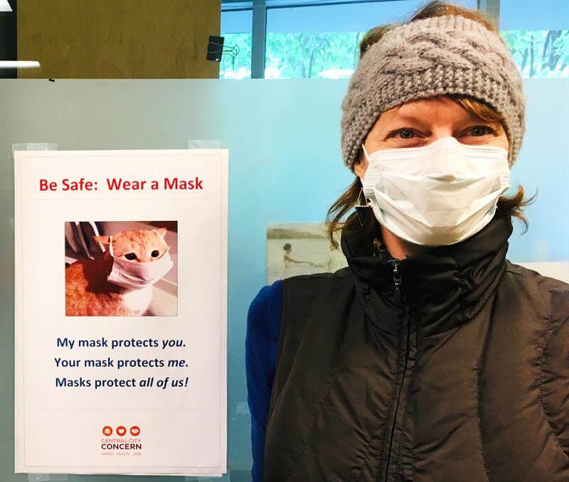 Person standing by sign encouraging other to protect one another by keeping their face mask on