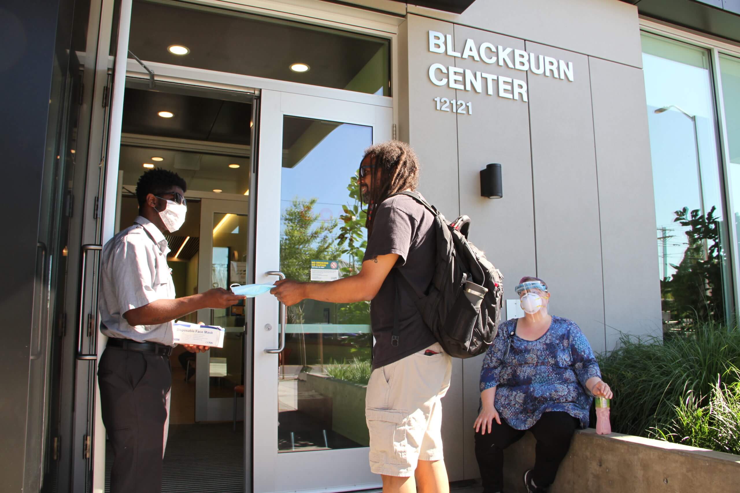Person holding door open for individual while also handing them a mask as they enter blackburn center building